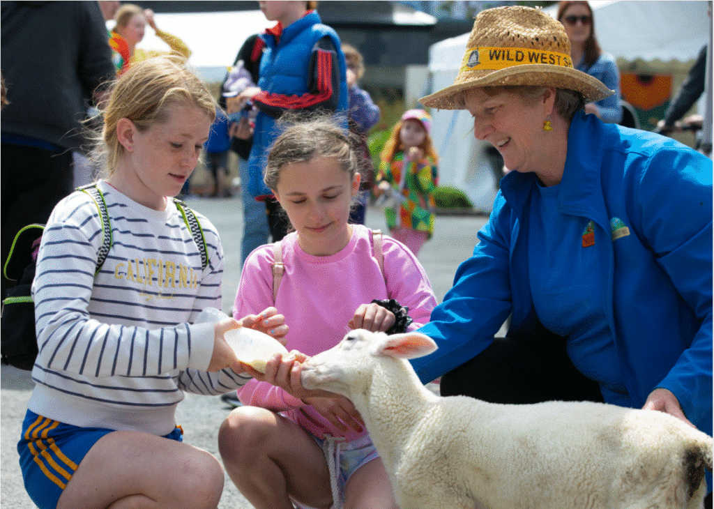 A woman gently pets a sheep while two children watch and smile nearby in a sunny outdoor setting.